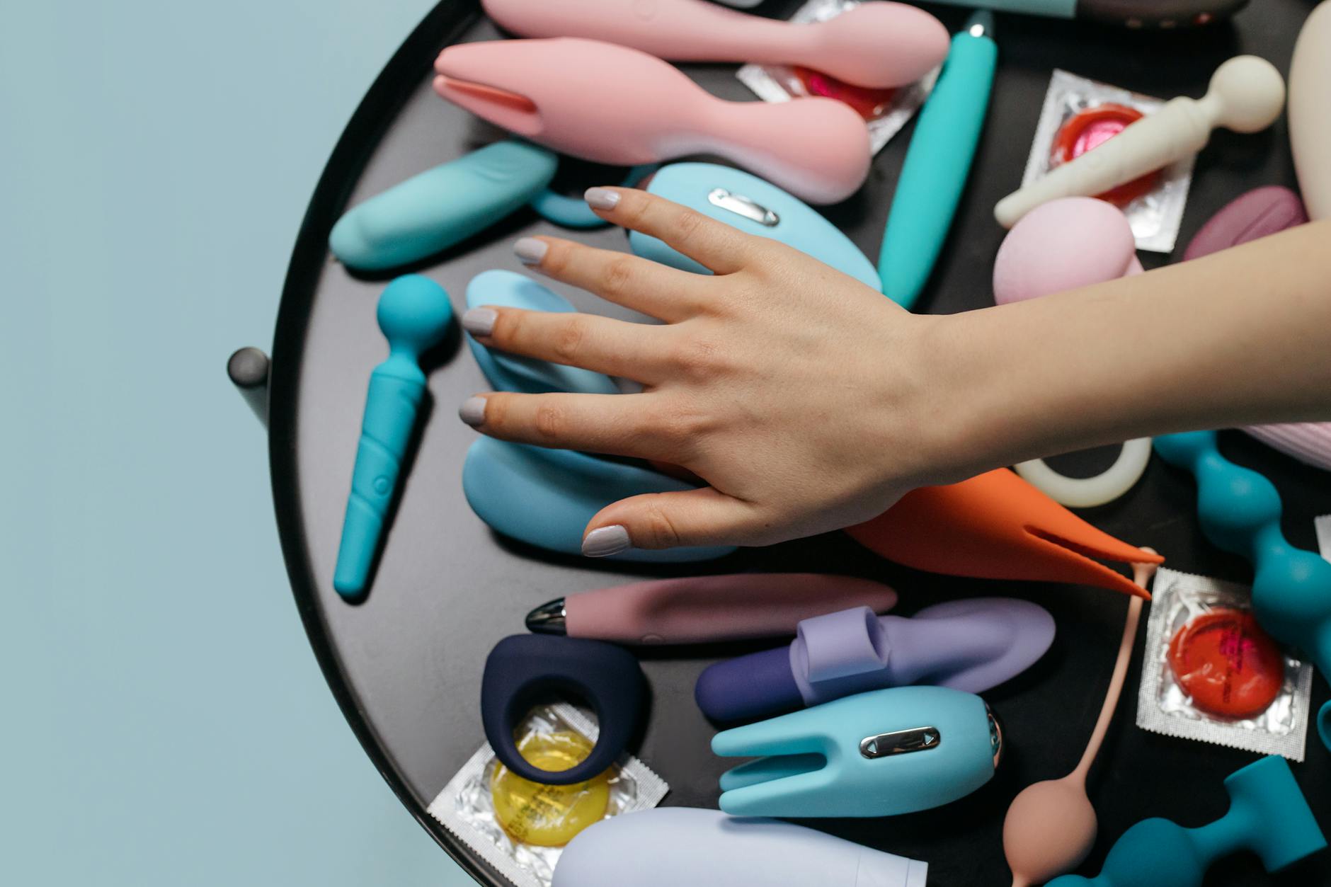 A hand reaching over a variety of colorful sex toys arranged on a table, including modern lemon vibrators.