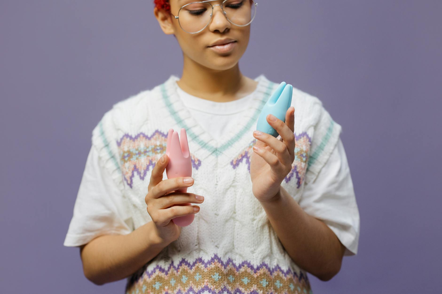 Woman with eyeglasses holding blue and pink silicone vibrators in a contemplative manner during pregnancy exploration.